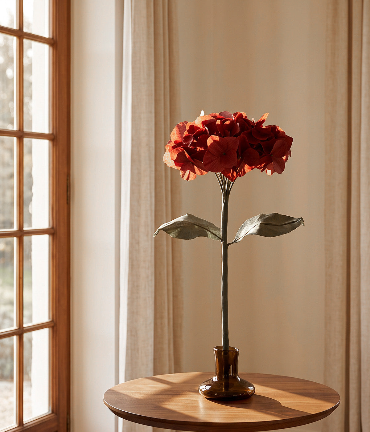 Burgundy paper hydrangea displayed on a mouth-blown glass stand beside a sunlit window